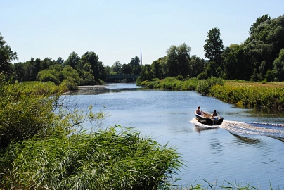 Zu sehen ist die Hamme, darauf schwimmend ein schmales Boot mit Motorantrieb, drumherum gibt es viele grüne Pflanzen.